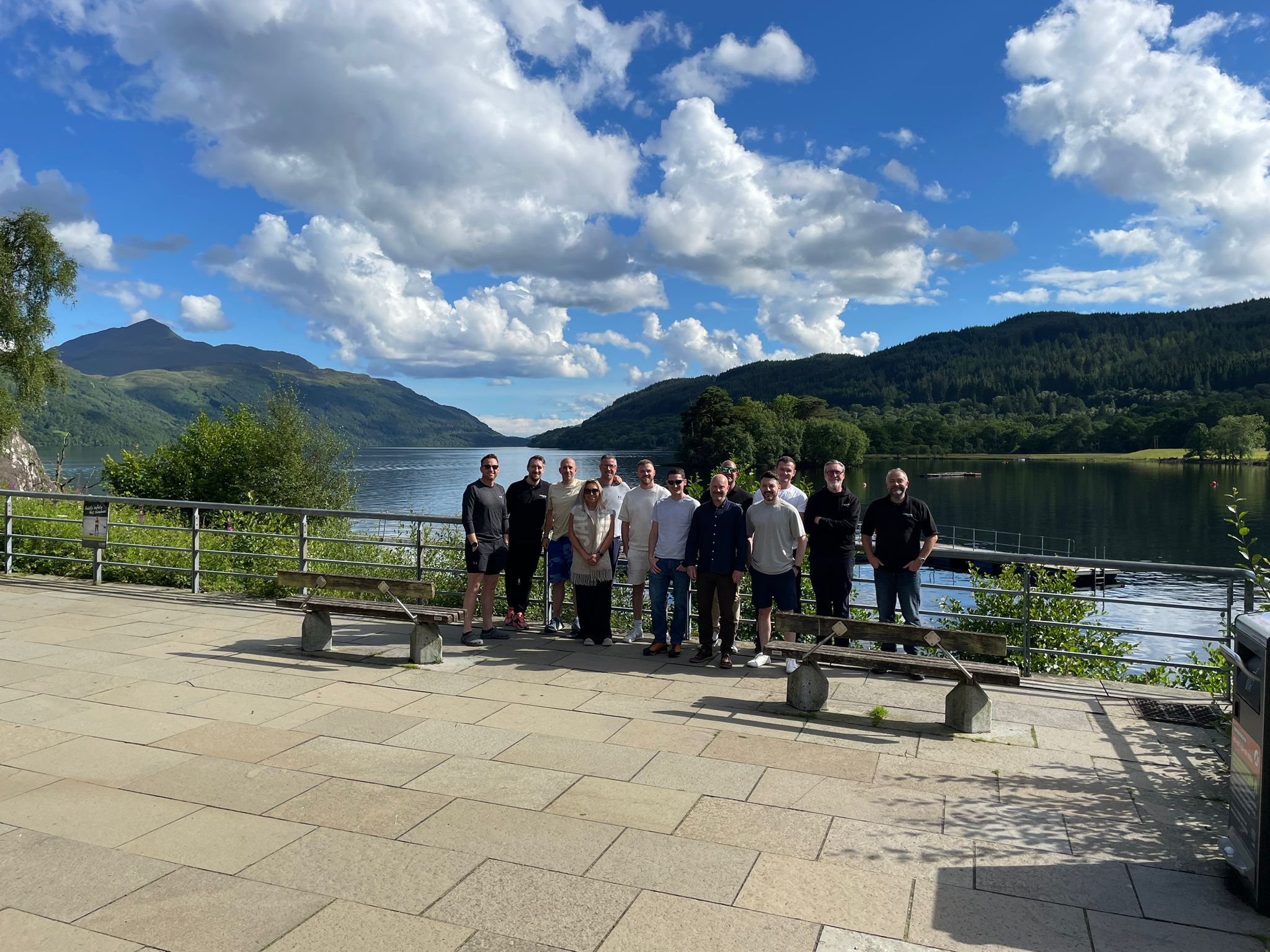 The team from Polypipe Building Products stand in front of Loch Lomond in Scotland. There is a blue sky with fluffy clouds and various green hills in the background.